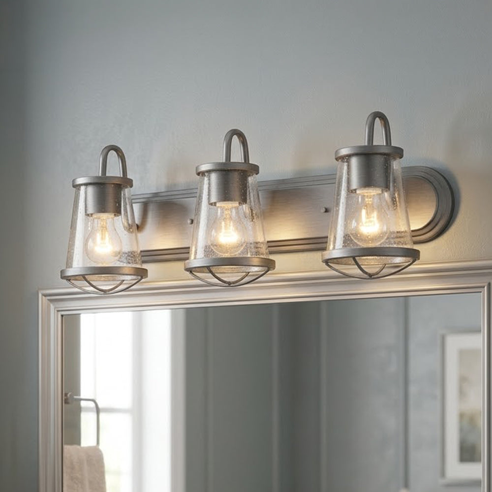 Three-light vanity wall sconce with clear glass shades and warm exposed bulbs, mounted above a mirror in a bathroom setting.