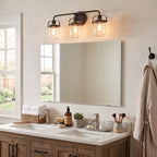 A three-light wall-mounted vanity fixture with clear glass shades and warm Edison bulbs, mounted above a rectangular mirror in a bright bathroom setting.