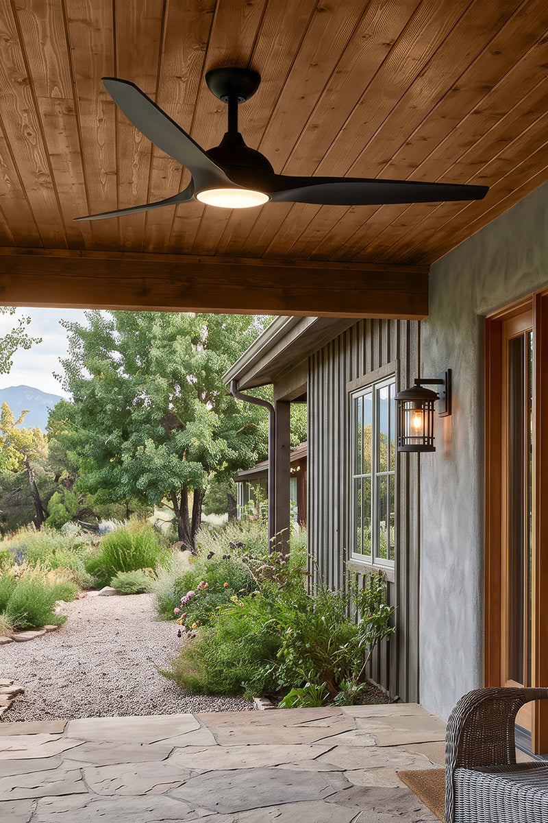 A black three-blade ceiling fan with an integrated light fixture hangs from a wooden covered porch. The porch has a stone tile floor, a rustic wall sconce, and a view of a landscaped garden and trees in the background.
